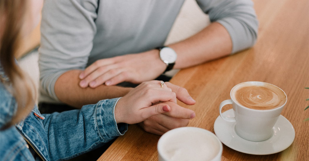 男性と女性がカフェで手をつないでいる写真　女性の手には婚約指輪が光っている